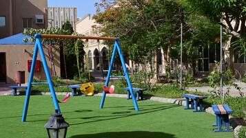 A playground with swings and benches, surrounded by greenery and buildings.