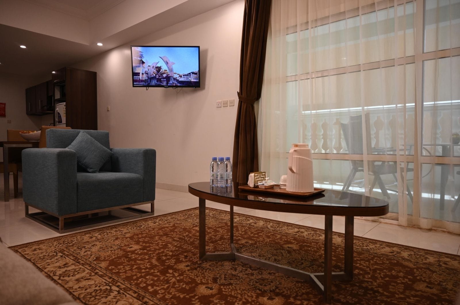 A modern hotel room with a blue armchair, a coffee table with amenities, a large window with curtains, and a flat-screen TV on the wall.