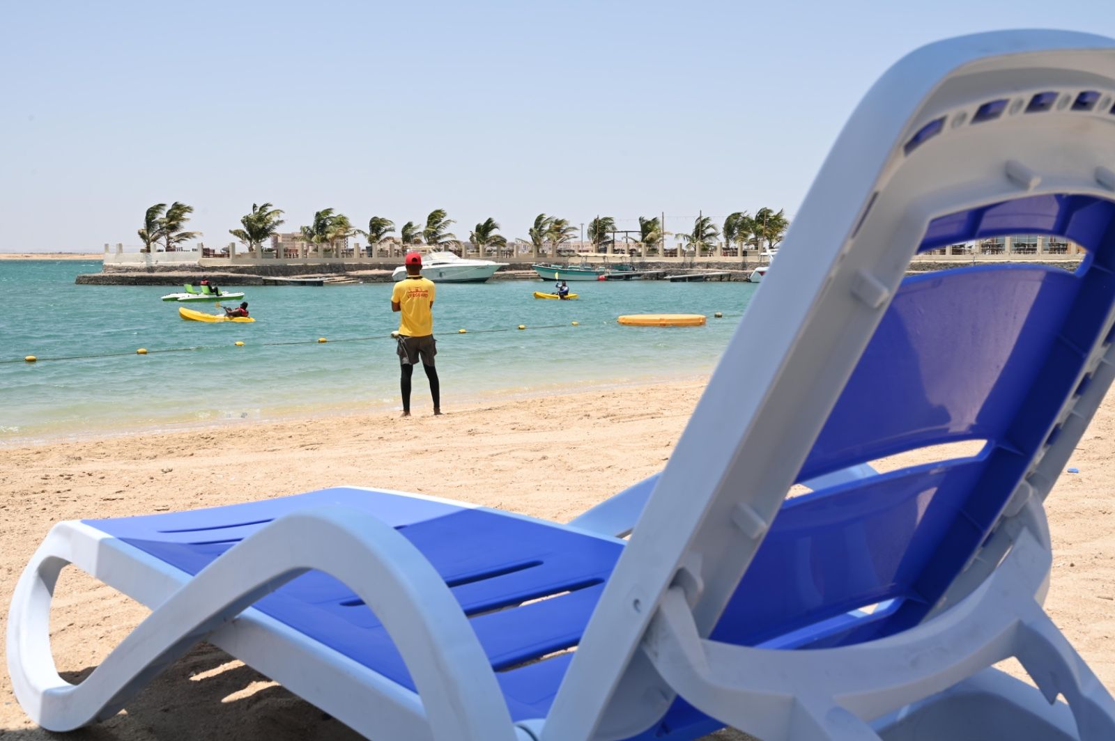 Una playa con una silla de lounge azul y blanca, una orilla arenosa y una persona con camisa amarilla junto al agua.