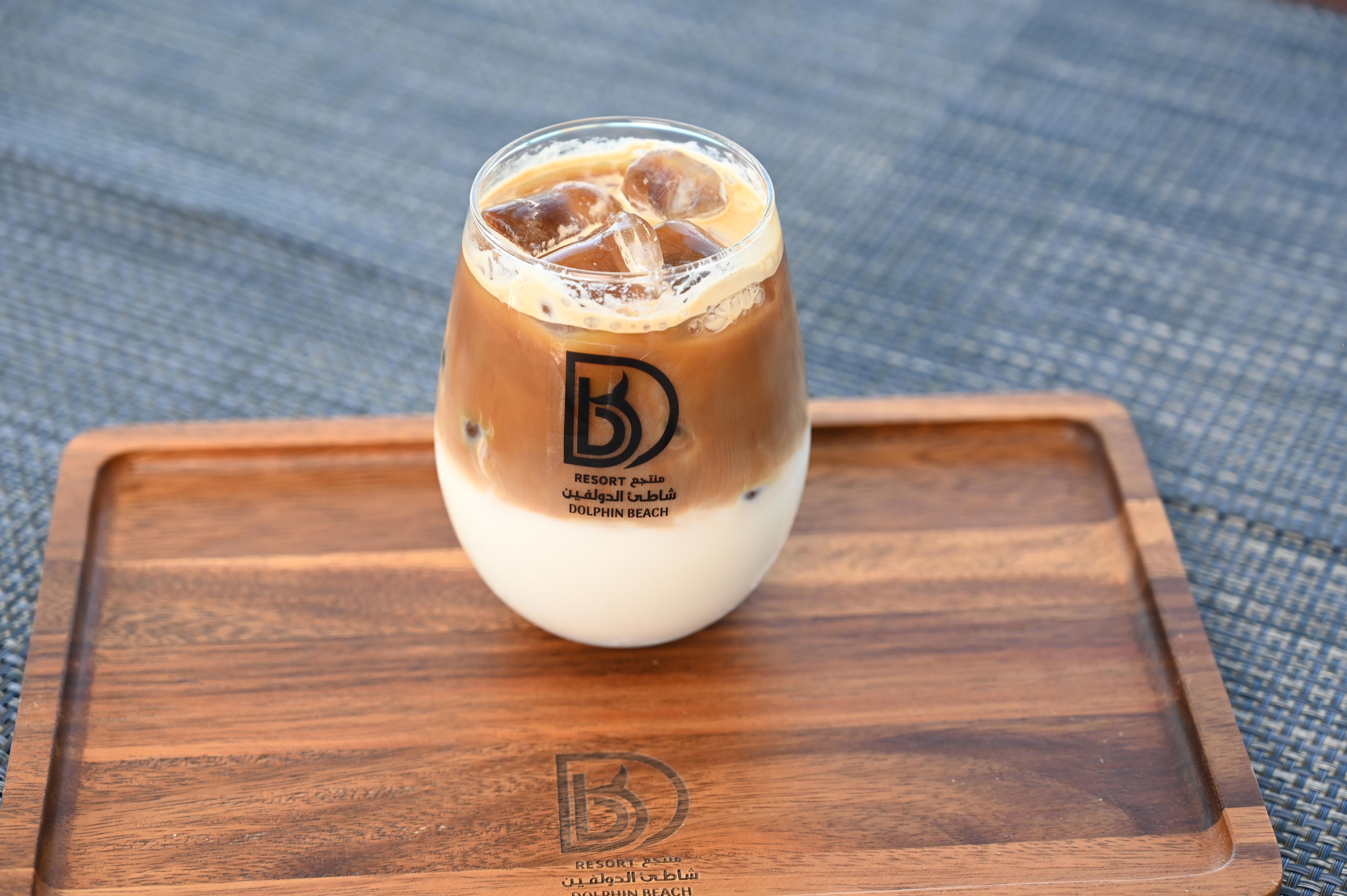 A glass of iced coffee with a logo on the side, placed on a wooden tray.