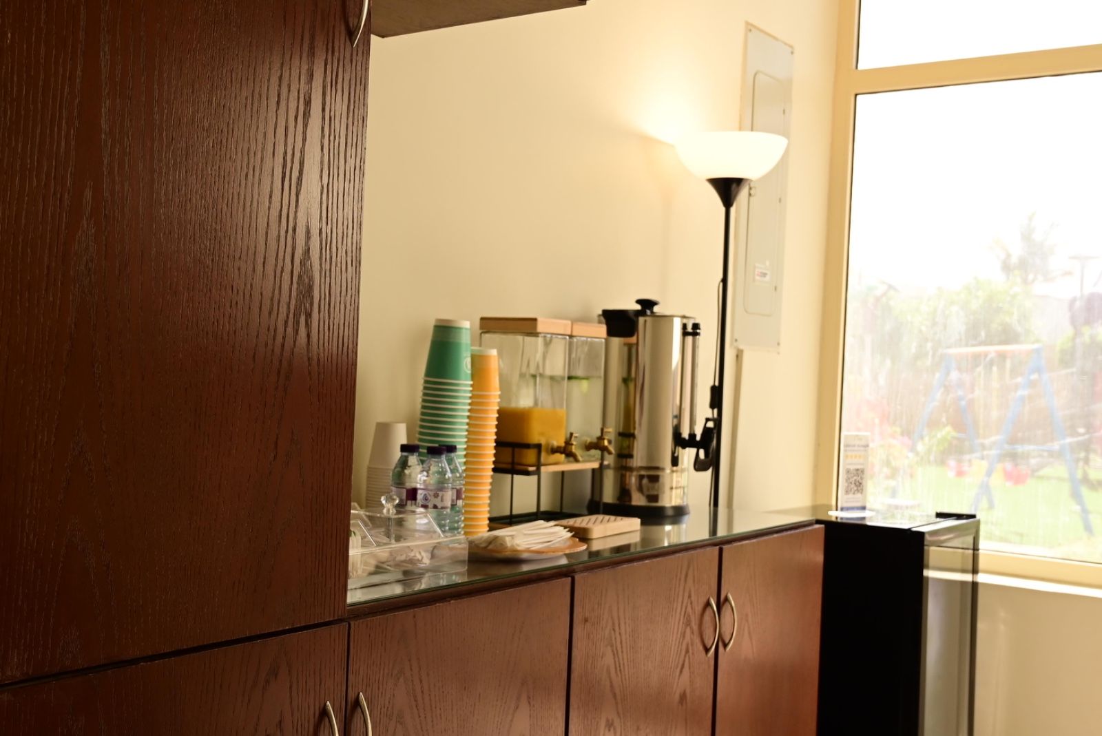 A kitchen counter with a coffee maker, stacked cups, and a window with a view of a garden.
