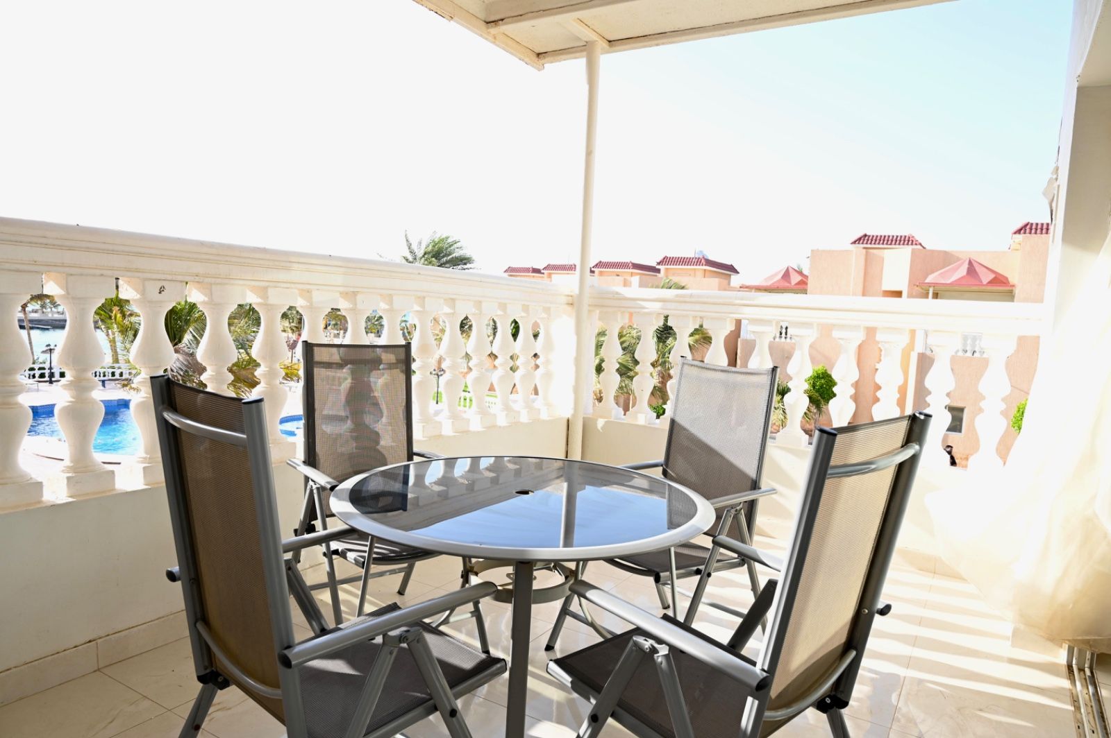 A round glass table with four chairs on a balcony with a white balustrade, overlooking a pool and buildings.