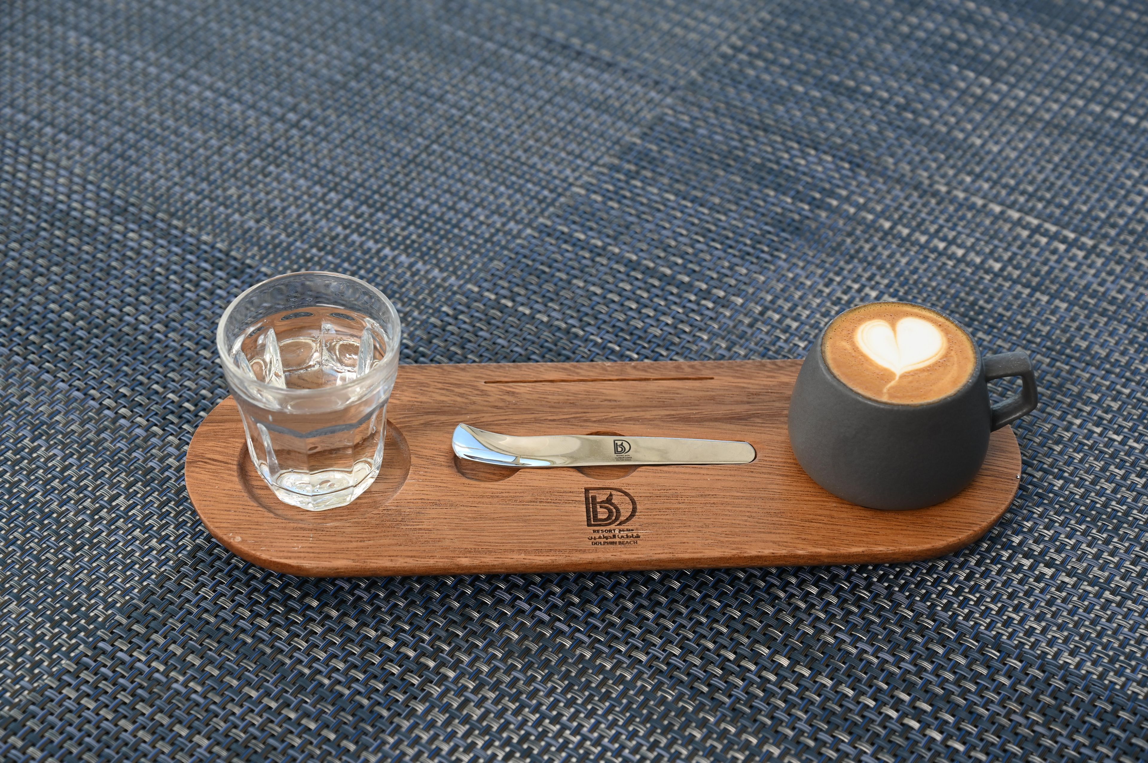 A wooden tray with a glass of water, a spoon, and a cup of coffee with a heart-shaped latte art on a woven surface.