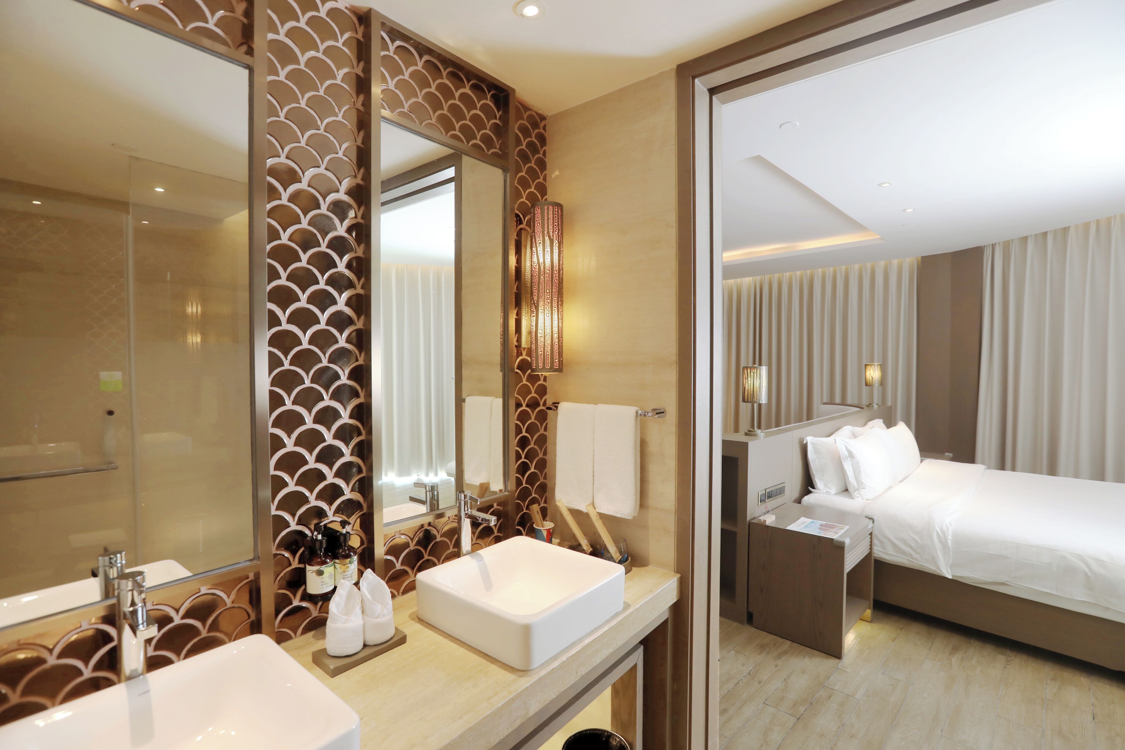 Habitación de hotel con un espejo grande, una mesita de noche, una cama y un baño con lavamanos y espejo.