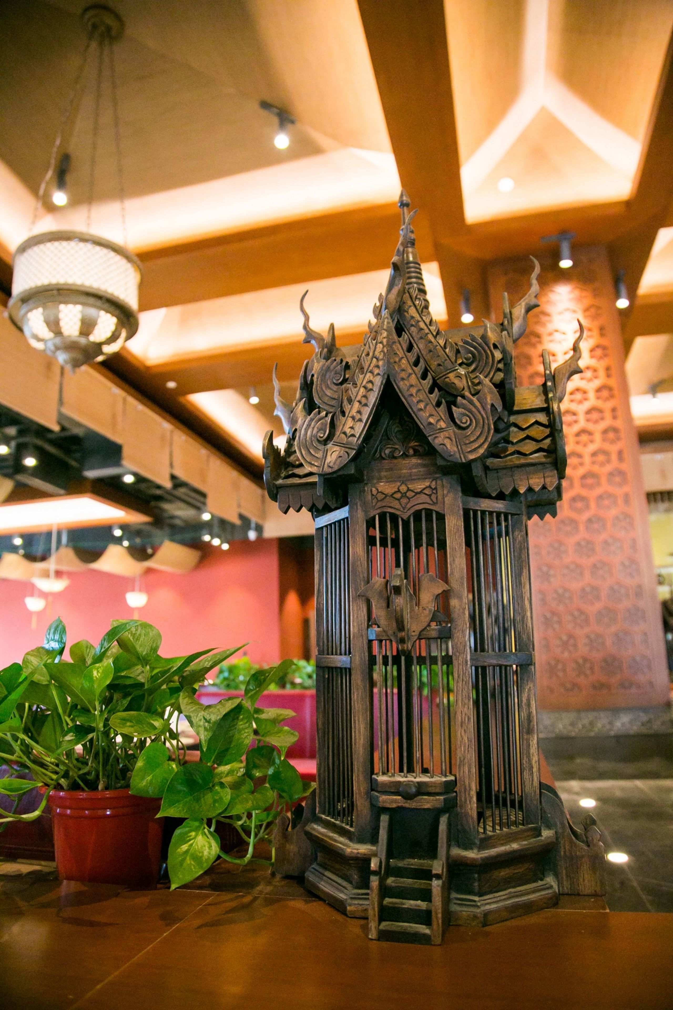Une structure traditionnelle en bois, ornée de sculptures complexes, peut-être un élément décoratif ou un petit sanctuaire, placée sur un plancher poli, avec des plantes en pot à proximité.