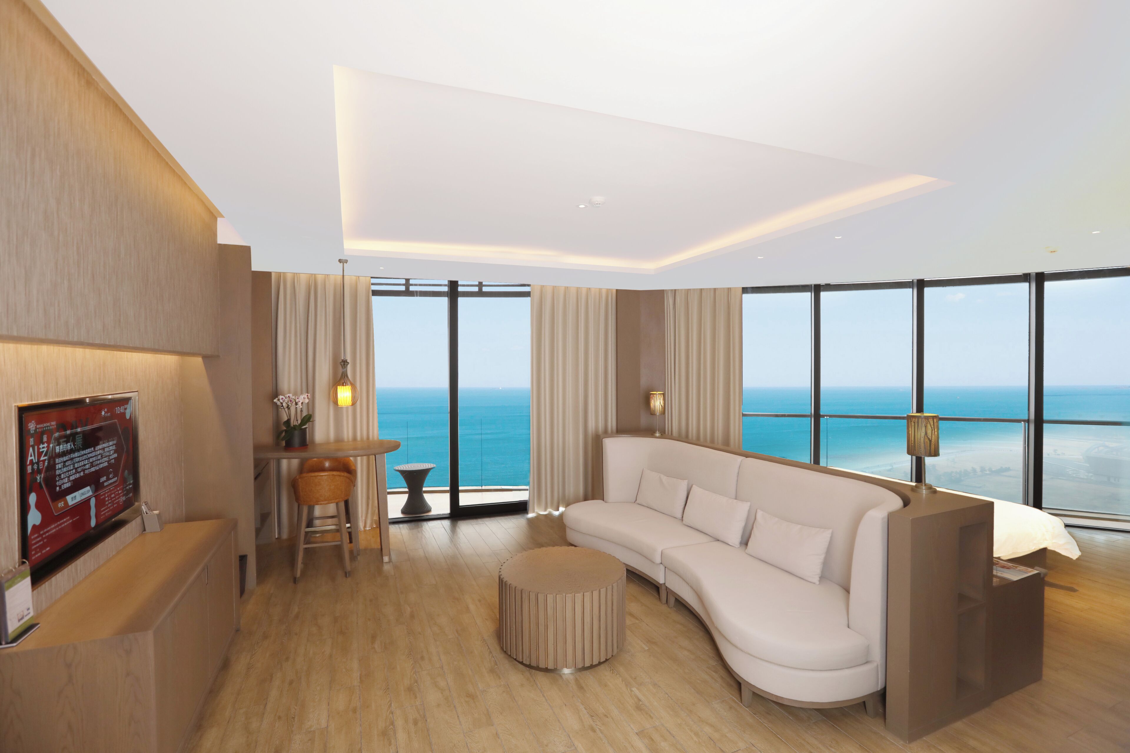 A modern hotel room with a large window, a sofa, a round ottoman, a desk, and a TV.