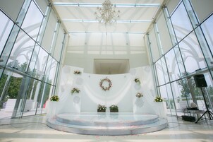 A glass-walled stage with a white backdrop, floral decorations, and a chandelier.