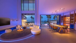A modern living room with a TV, seating, and a dining area with a view of the beach.