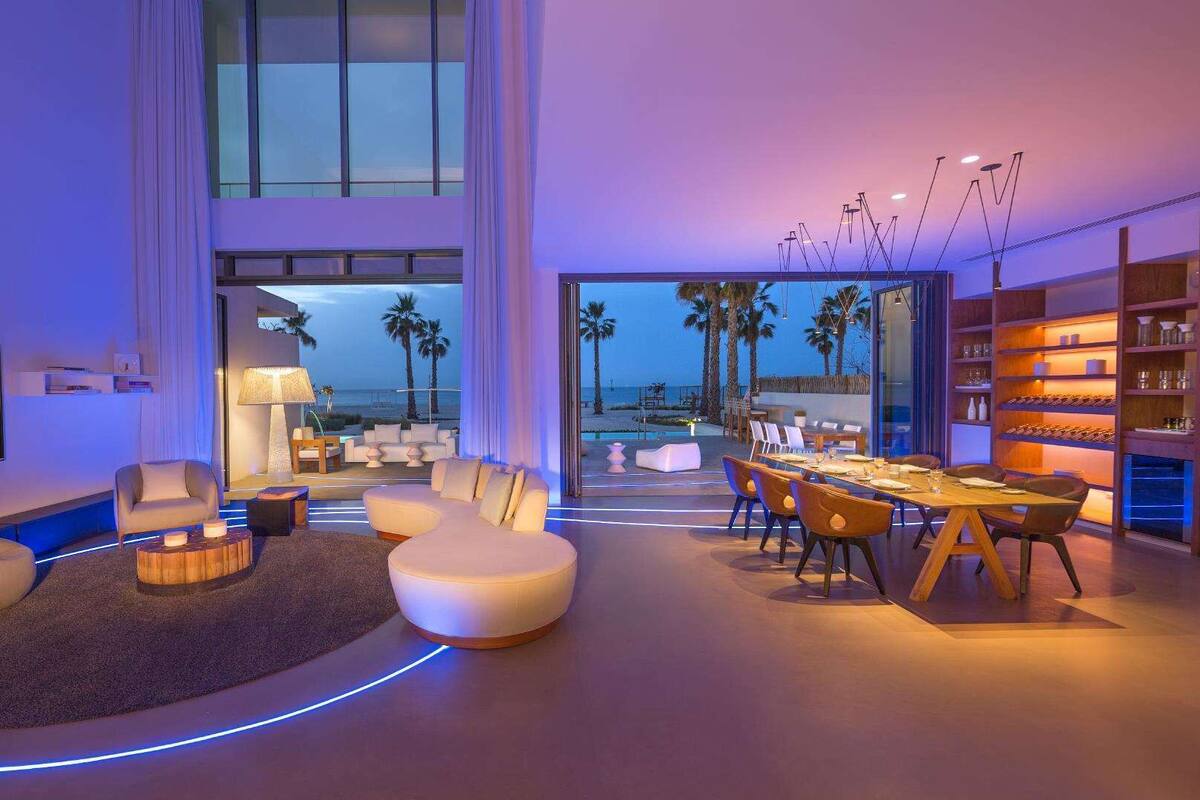 A modern living room with a TV, seating, and a dining area with a view of the beach.