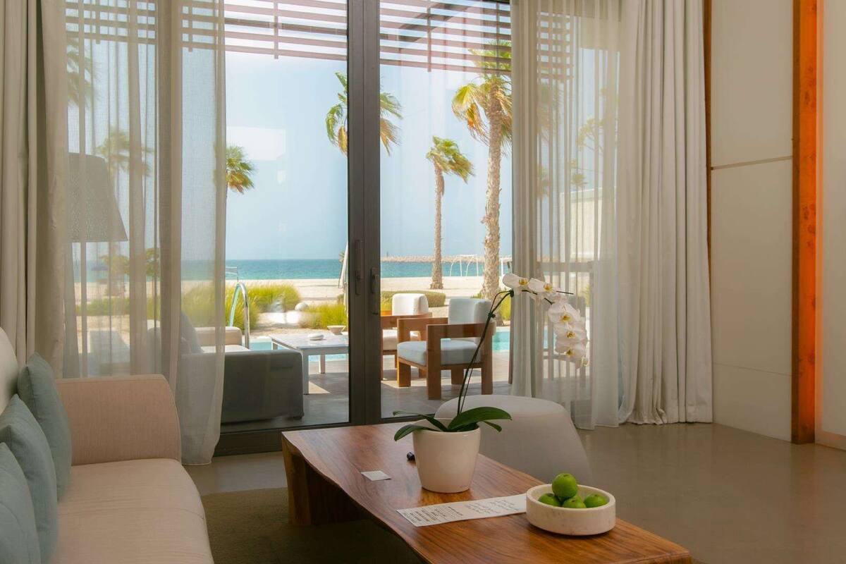 A modern living room with a view of the beach, a wooden coffee table with a plant and fruit bowl, and a sofa with cushions.