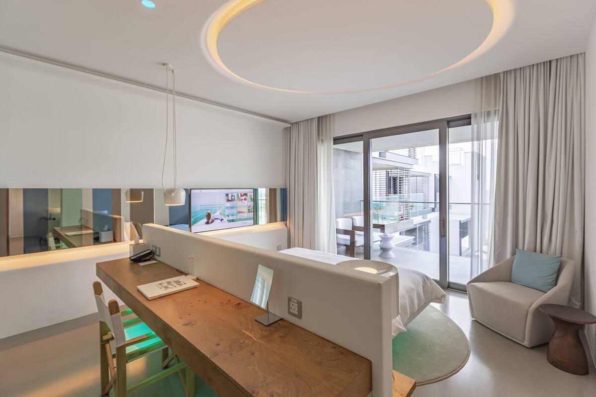 A modern hotel room with a wooden desk, a large window with curtains, a sofa, and a view of the city.