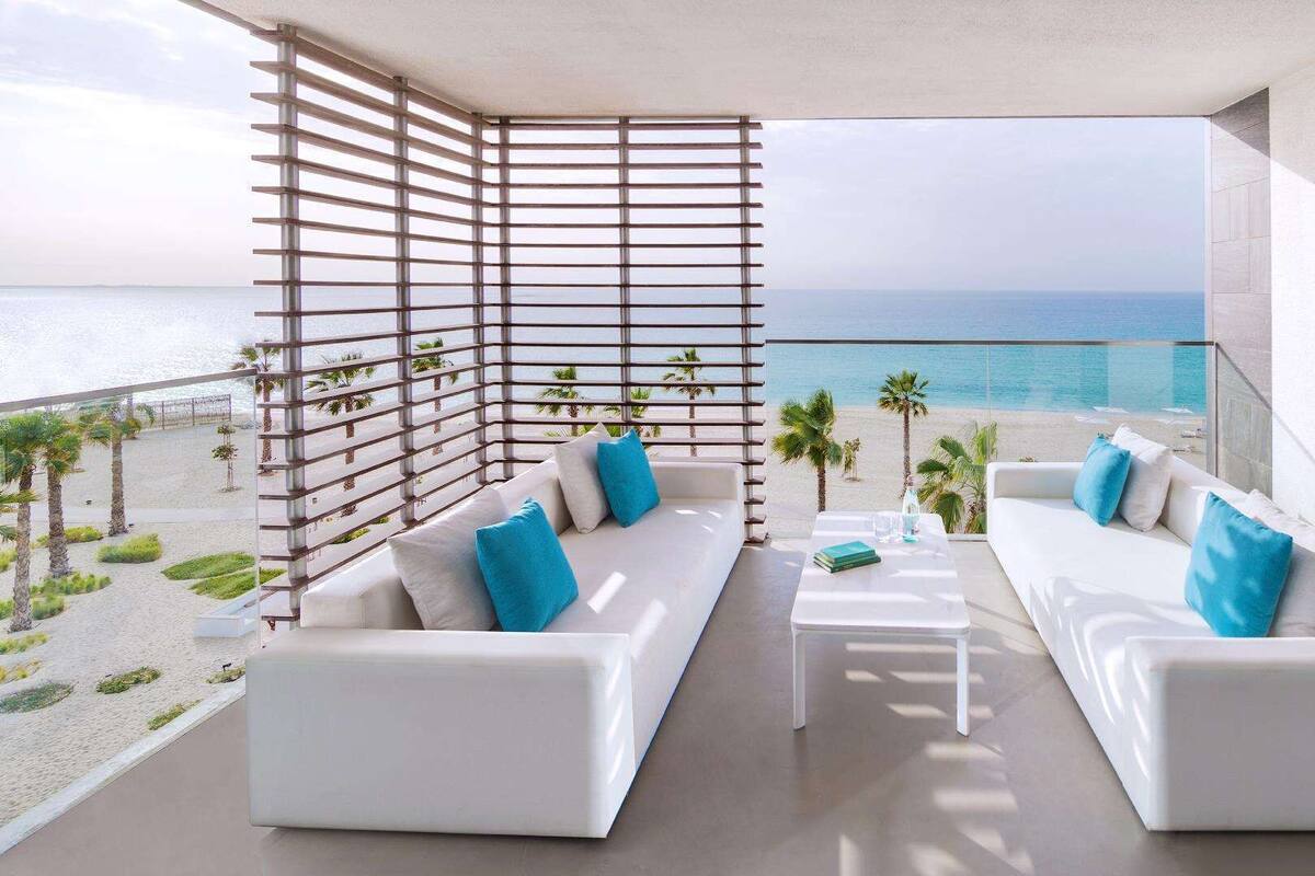 A modern balcony with white sofas, blue cushions, and a view of the beach and ocean.