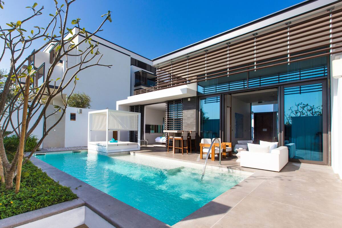 A modern outdoor pool area with a clear blue pool, white outdoor furniture, and a contemporary building with large windows.