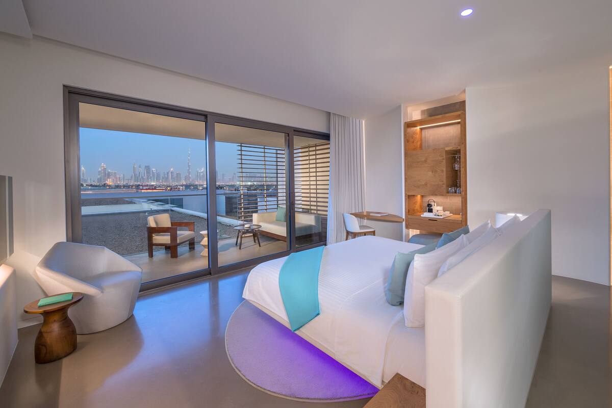 A modern hotel room with a large bed, a seating area with a view of the city, and a small desk with a chair.