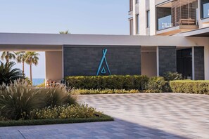 A modern building with a geometric logo on the facade, a paved entrance, and a landscaped area with palm trees and shrubs.