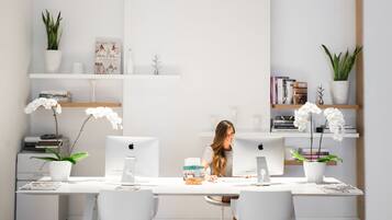 Un espace de bureau moderne et minimaliste, avec des meubles blancs, notamment un bureau et deux chaises. On trouve deux ordinateurs Apple iMac sur le bureau, et la pièce est dotée d’étagères blanches où sont disposés des objets décoratifs et des plantes.