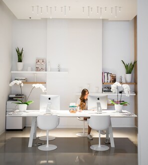 A modern, minimalist office space with white furniture, including a desk and two chairs. There are two Apple iMac computers on the desk, and the room has white shelving with decorative items and plants.