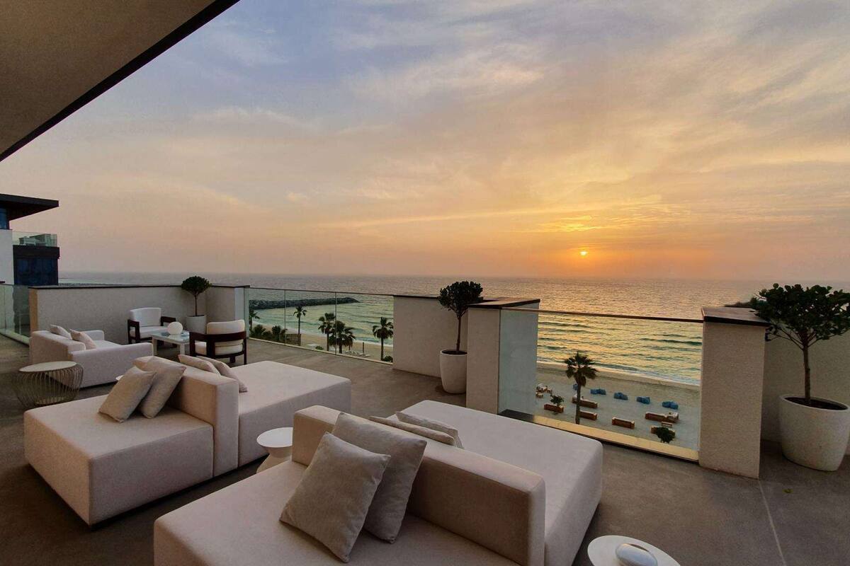 A modern outdoor seating area with sofas and a table, overlooking a beach and ocean at sunset.