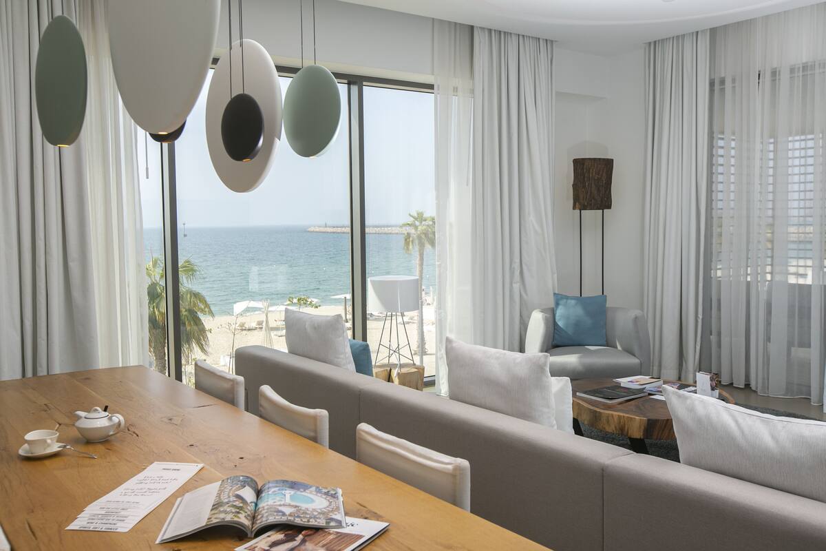A modern hotel room with a wooden dining table, a sofa, and a view of the sea.