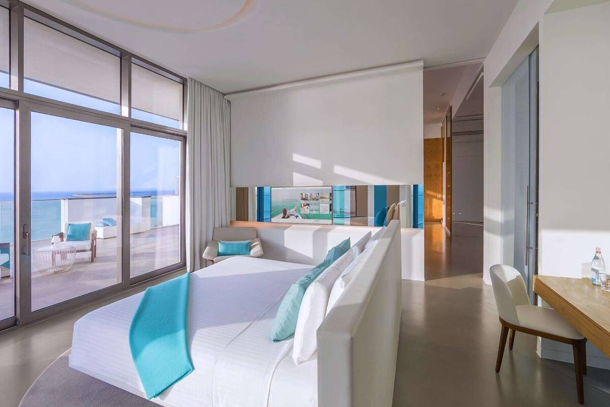 A modern hotel room with a large balcony, a bed with white bedding and blue accents, a small sitting area, and a view of the ocean.