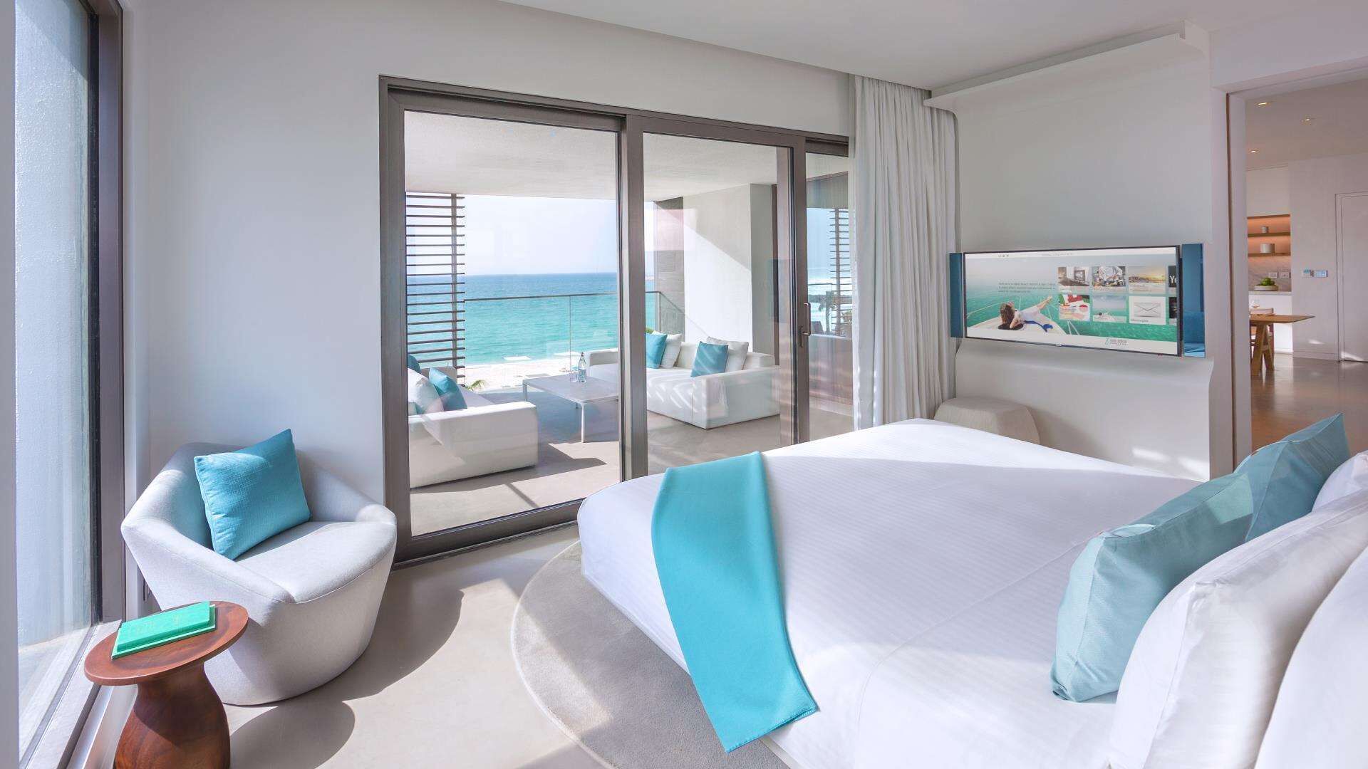 Habitación de hotel con cama, una silla, una mesita y un ventanal con vista al océano.