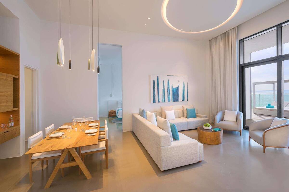 A modern living room with a wooden dining table set for four, a white sofa with blue cushions, and a large window offering a view of the sea.