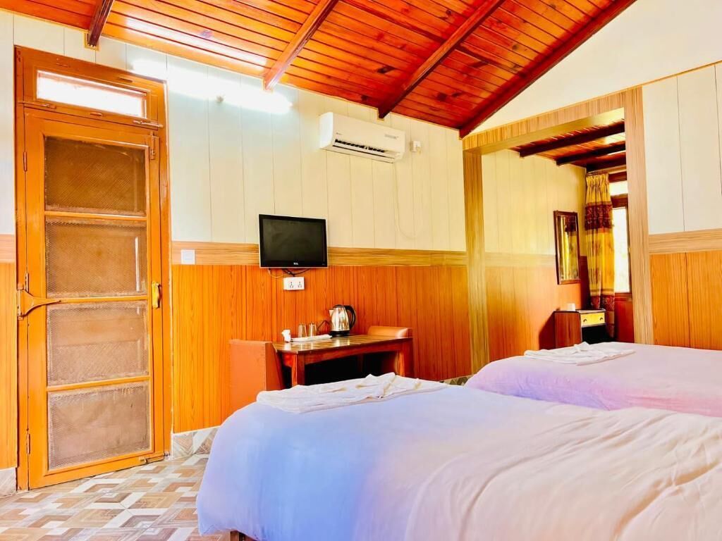 A hotel room with two beds, a television, a desk with a kettle, and an air conditioning unit.