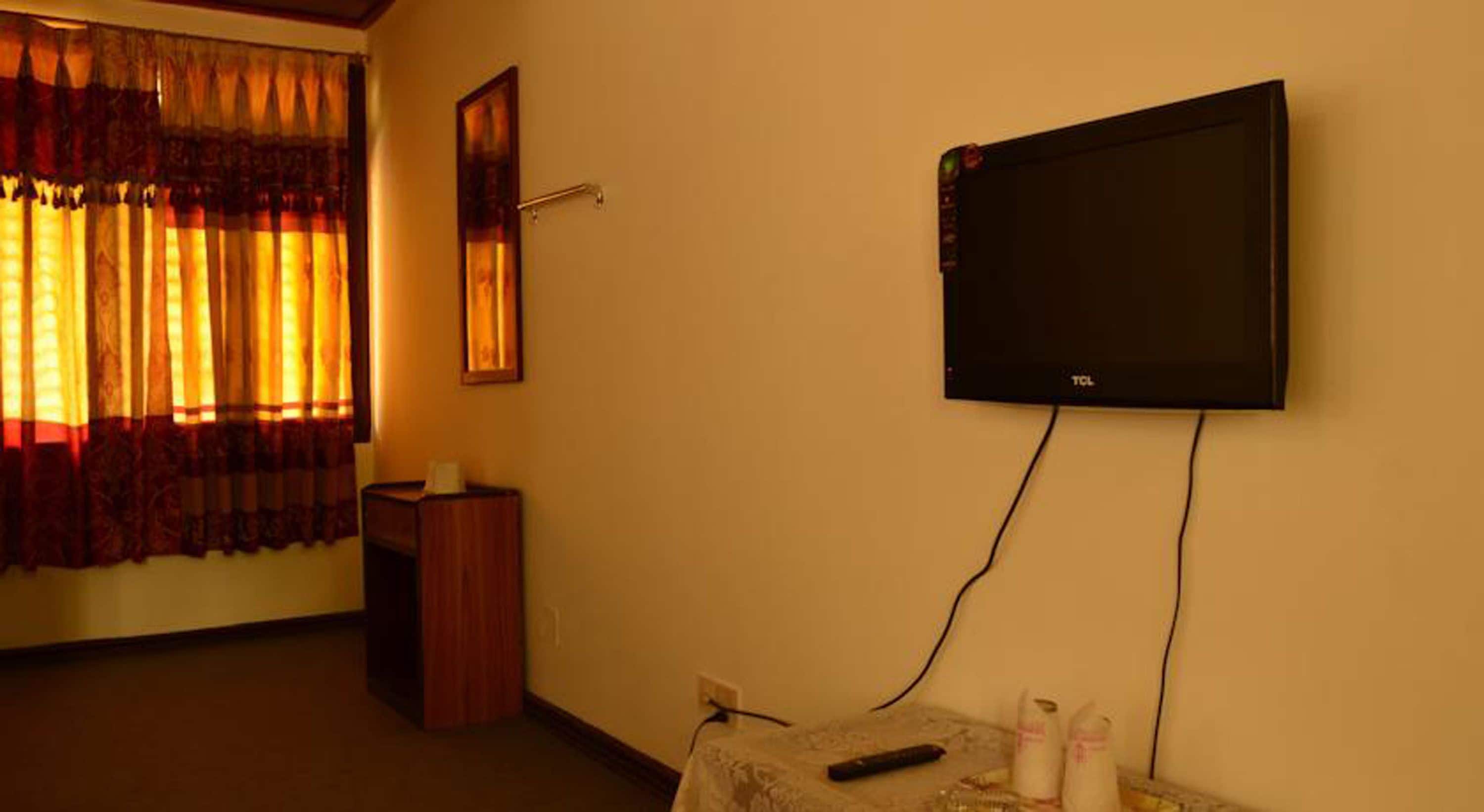 A room with a mounted television, a small table with remote controls, and a window with curtains.