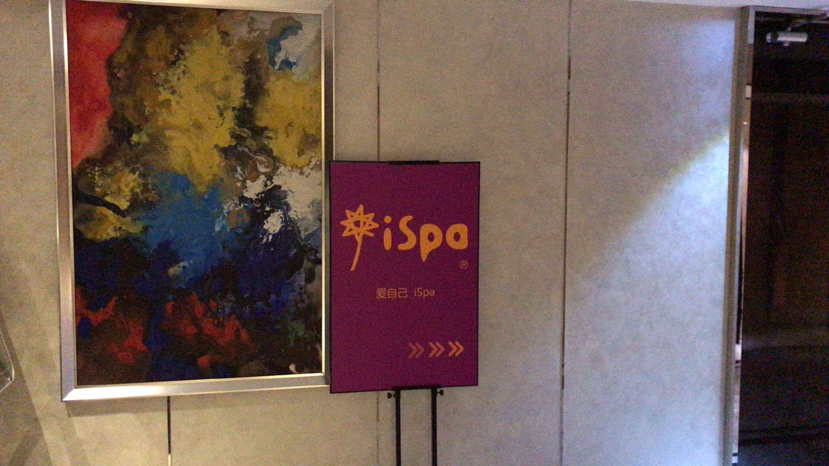 A purple sign with the text "iSpa" and Chinese characters, mounted on a stand against a wall with a colorful abstract painting.