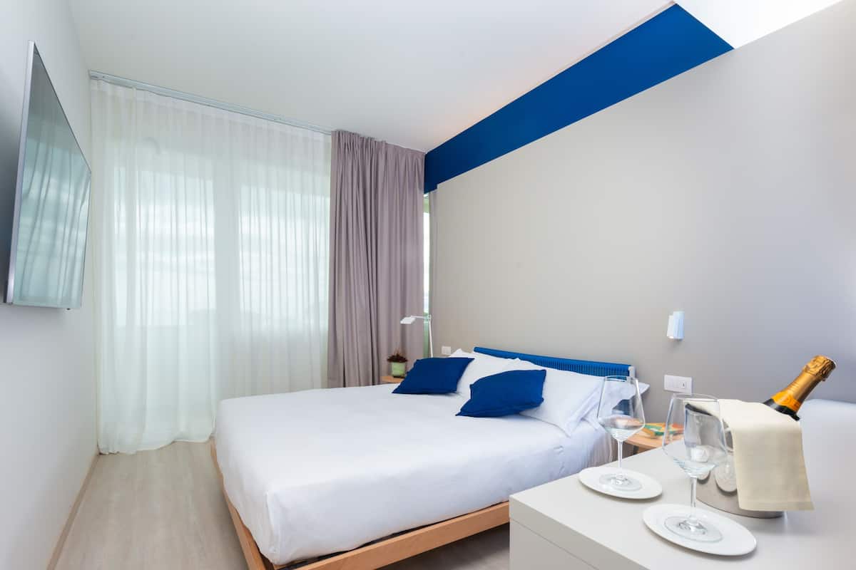 A modern hotel room with a large bed, a bedside table with a bottle of champagne, and a small table with two glasses.