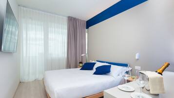 Habitación de hotel moderna con una cama grande, una mesita de noche con una botella de champán y una pequeña mesa con dos copas.