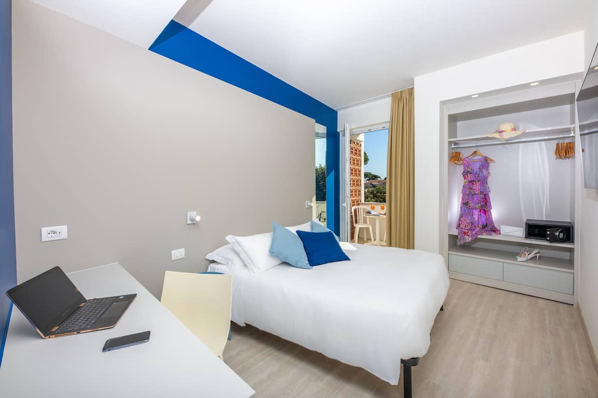 A hotel room with a bed, a desk with a laptop, a phone, and a wardrobe with clothes.