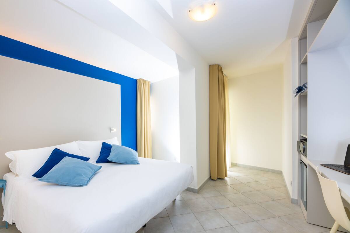 A modern hotel room with a bed, a desk, and a kitchenette.