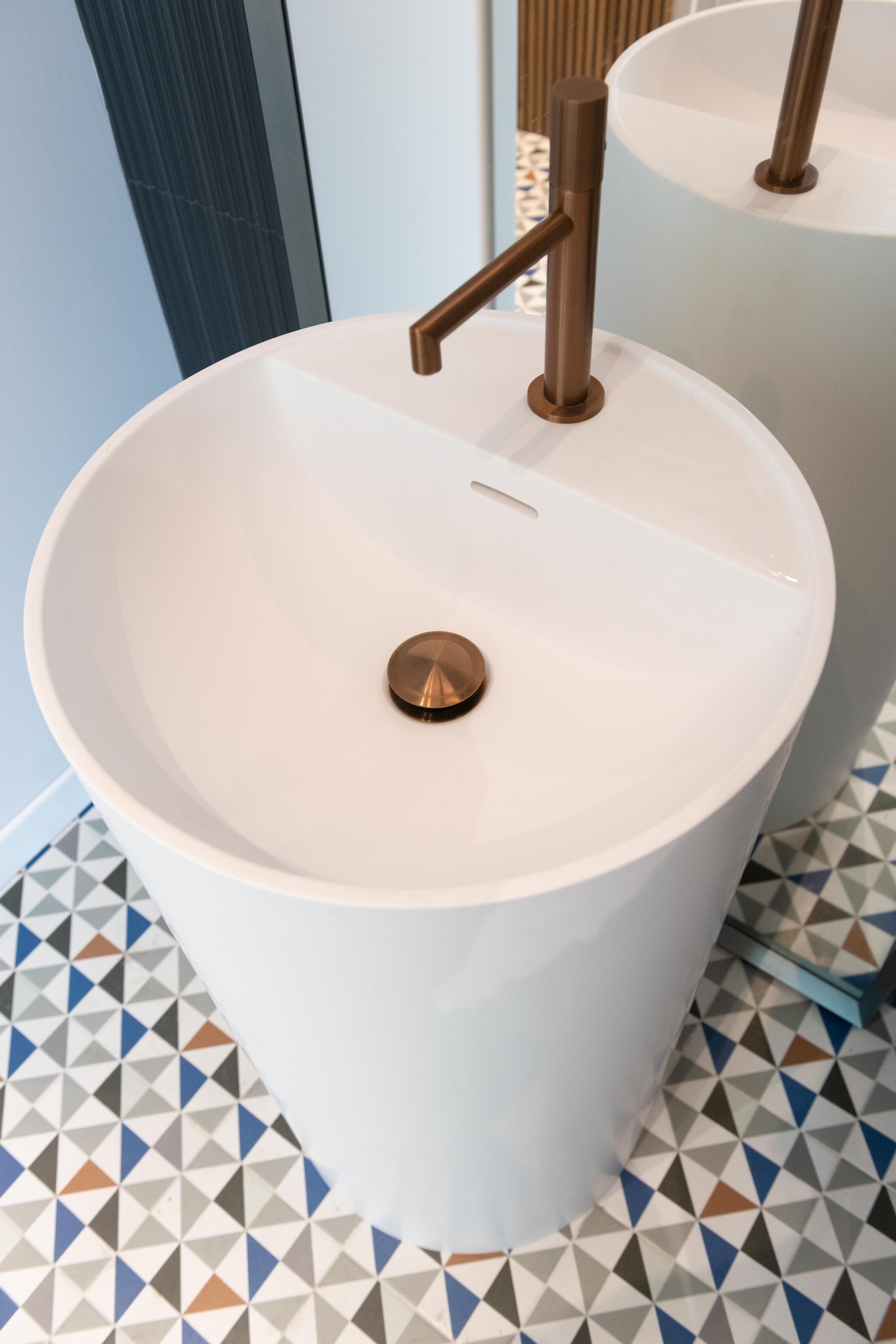 A white pedestal sink with a copper faucet and drain, set on a tiled floor with a geometric pattern.