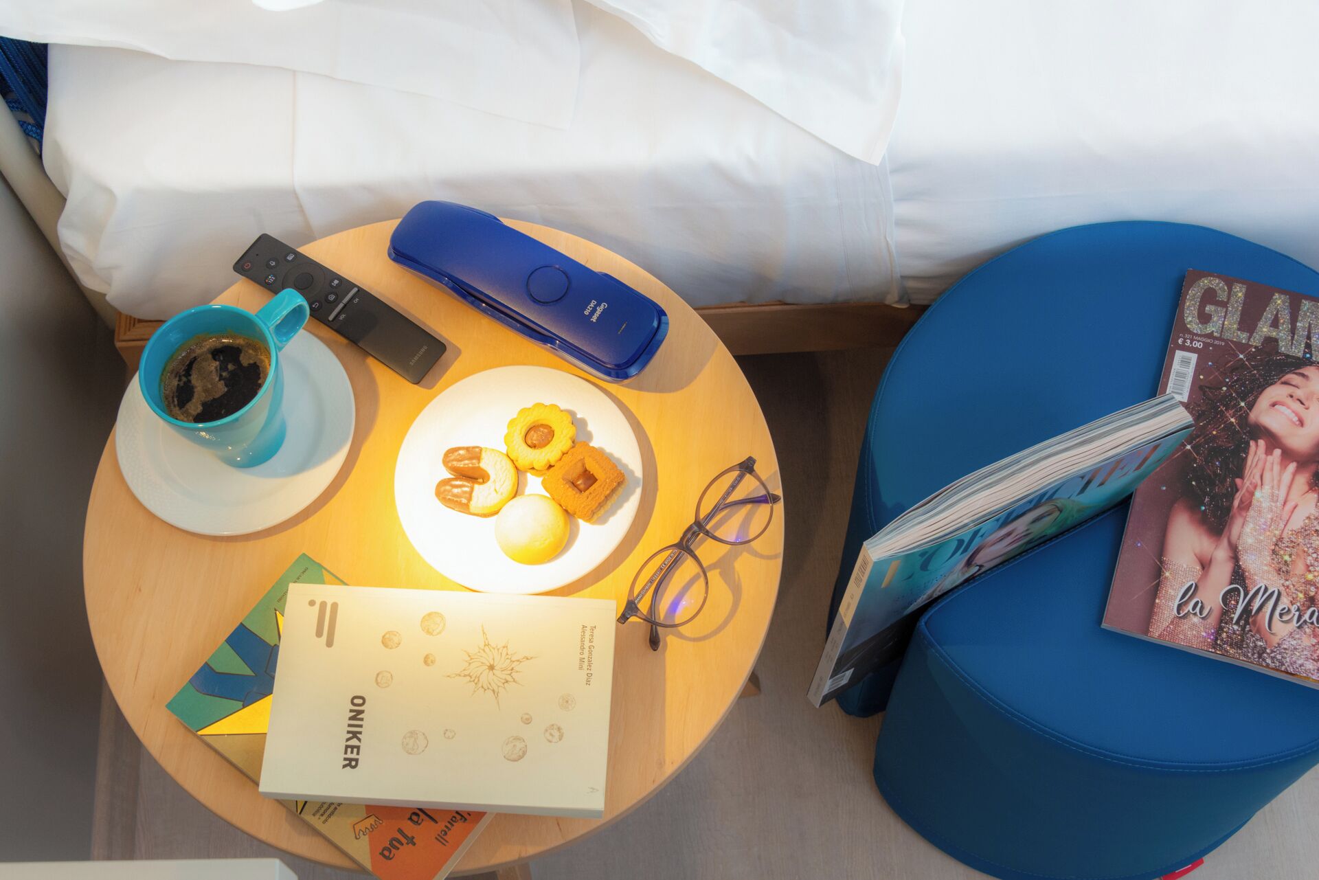 A round wooden table with a remote control, a mug, a plate with food, glasses, and a magazine.