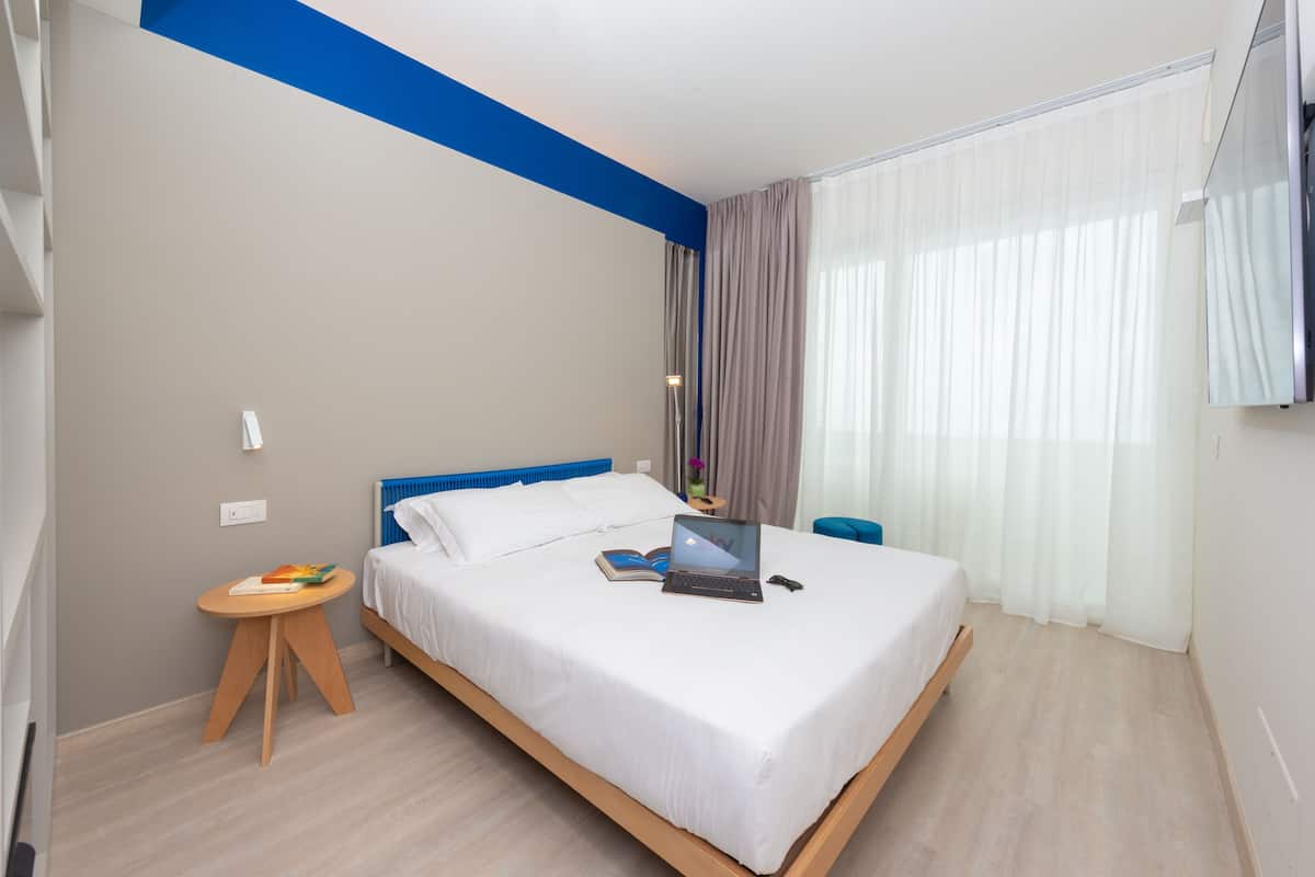 A modern hotel room with a large bed, a laptop on the bed, a small wooden side table, and a view of the outdoors through sheer curtains.