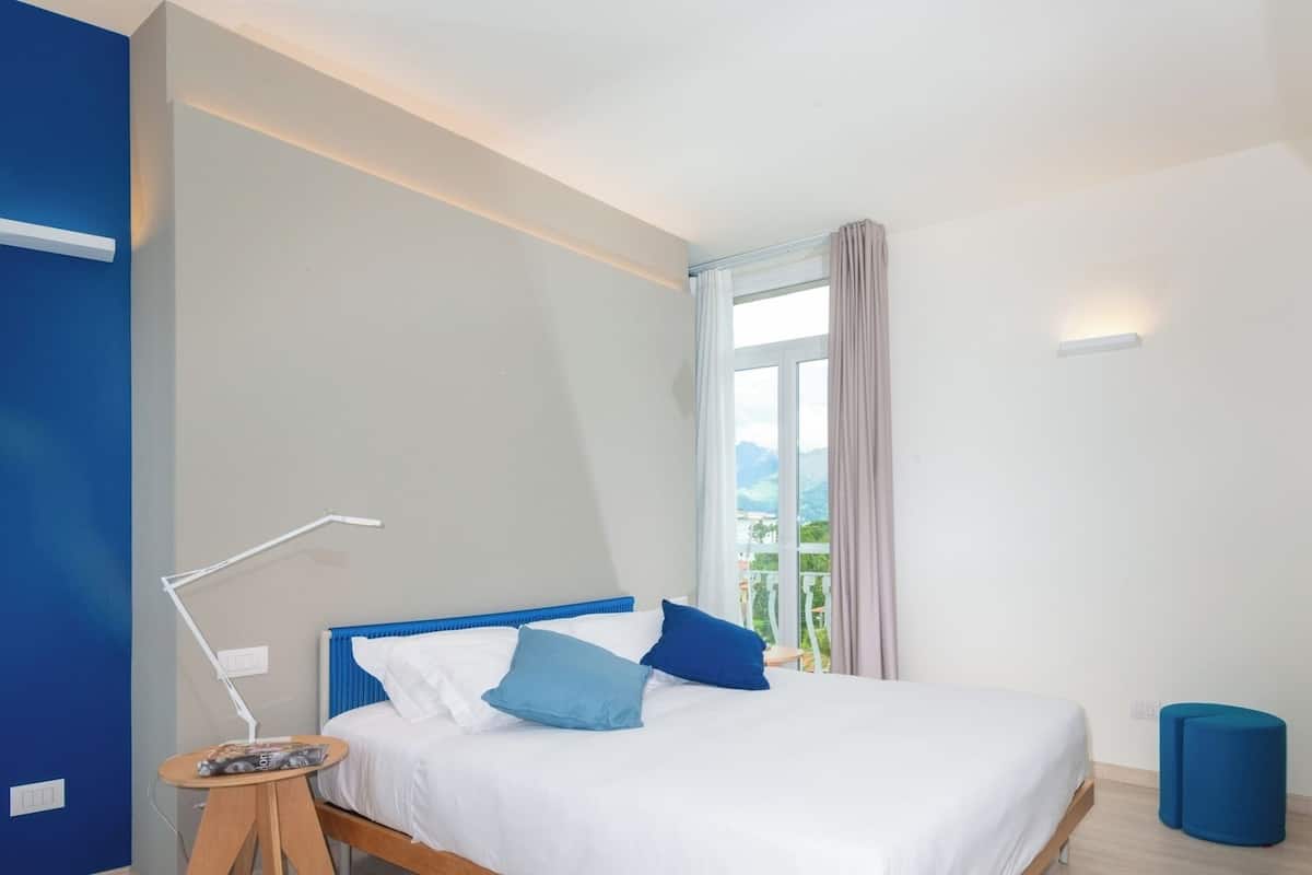 A modern hotel room with a bed, a small table, a lamp, and a view of the outdoors.