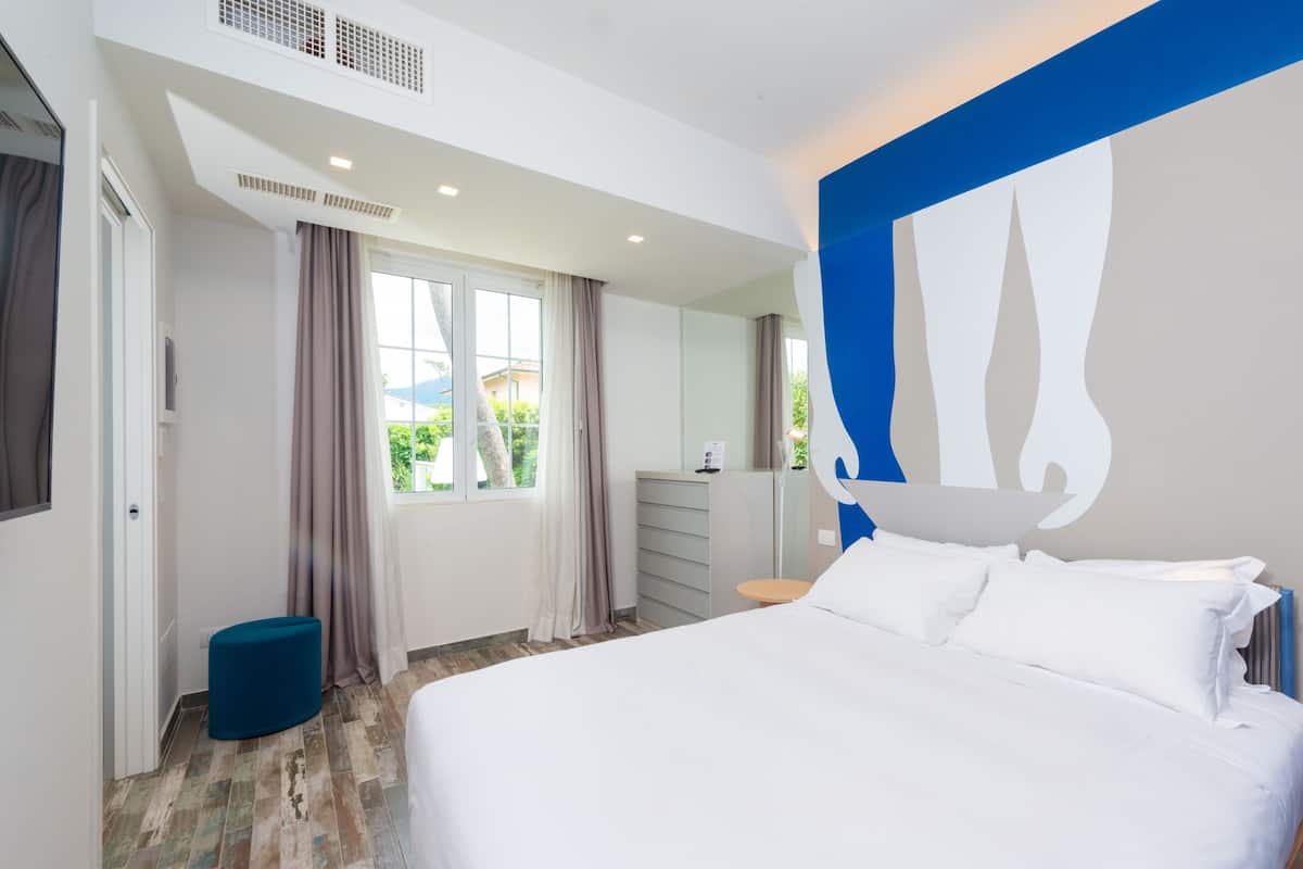 A modern hotel room with a large bed, a small round table, and a view of the outdoors through a window.