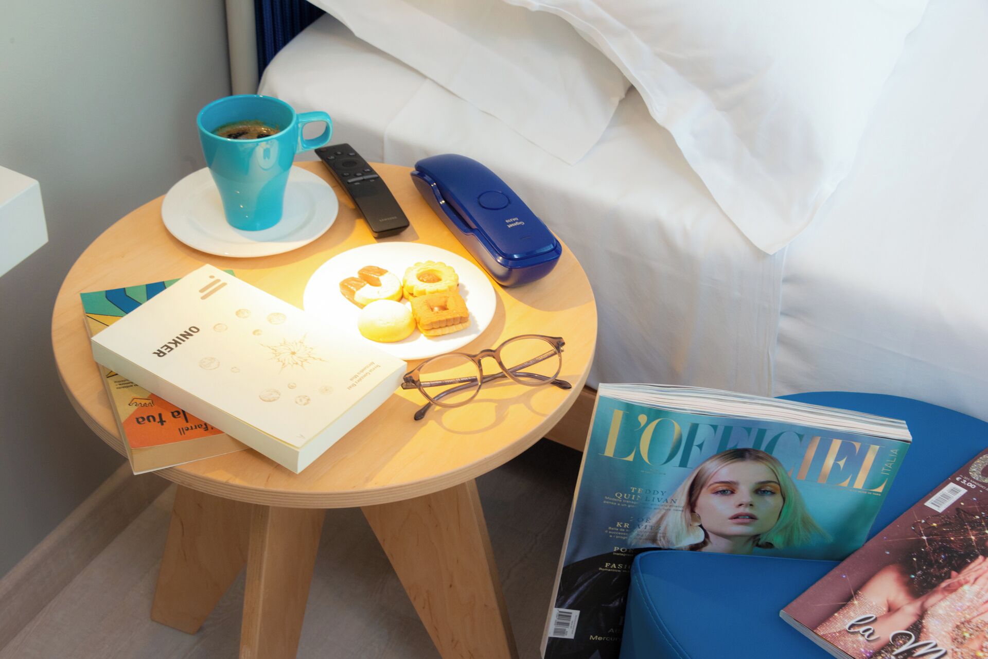 A small round wooden table with a book, a pair of glasses, a remote control, a blue mug, and a plate with pastries.