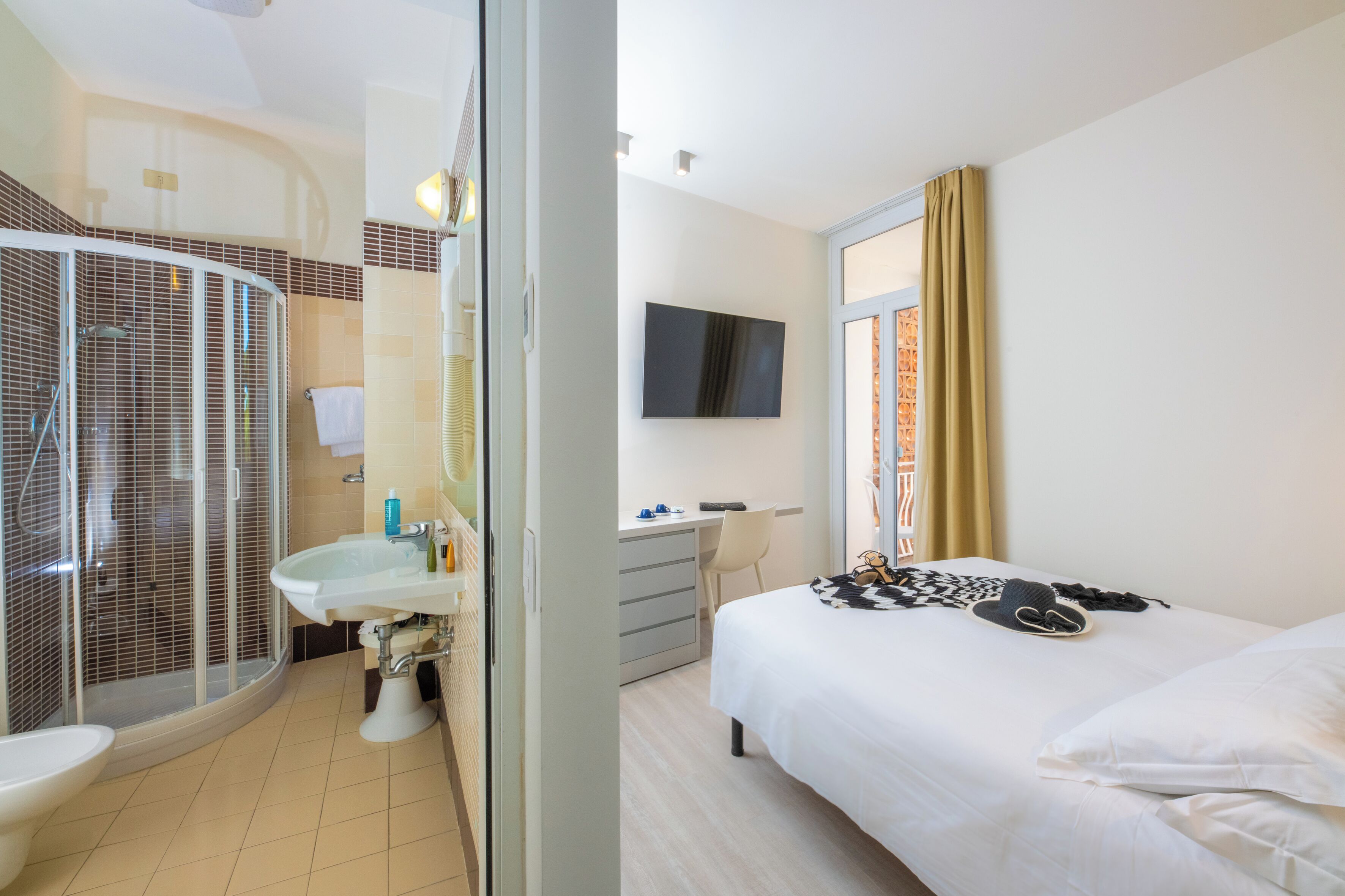 Habitación de hotel con ducha, lavamanos, cama, escritorio y televisor.