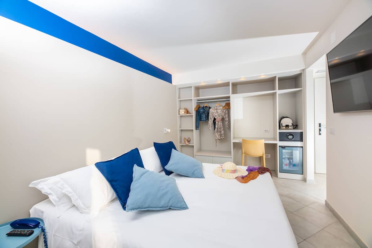 A modern hotel room with a neatly made bed, a wardrobe with hanging clothes, a small kitchenette, and a TV mounted on the wall.