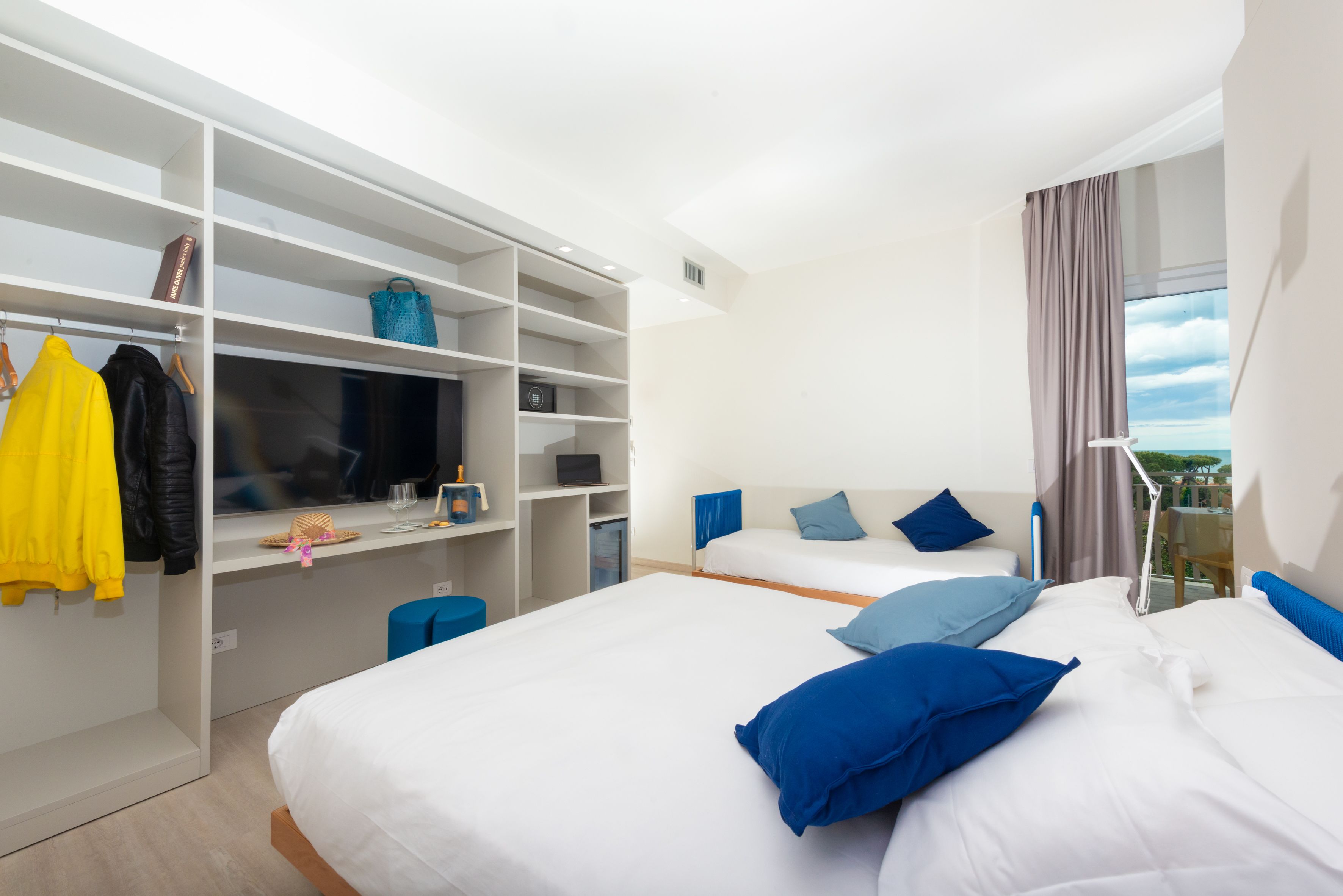 A hotel room with two beds, a wardrobe, a desk, and a view of the outdoors.