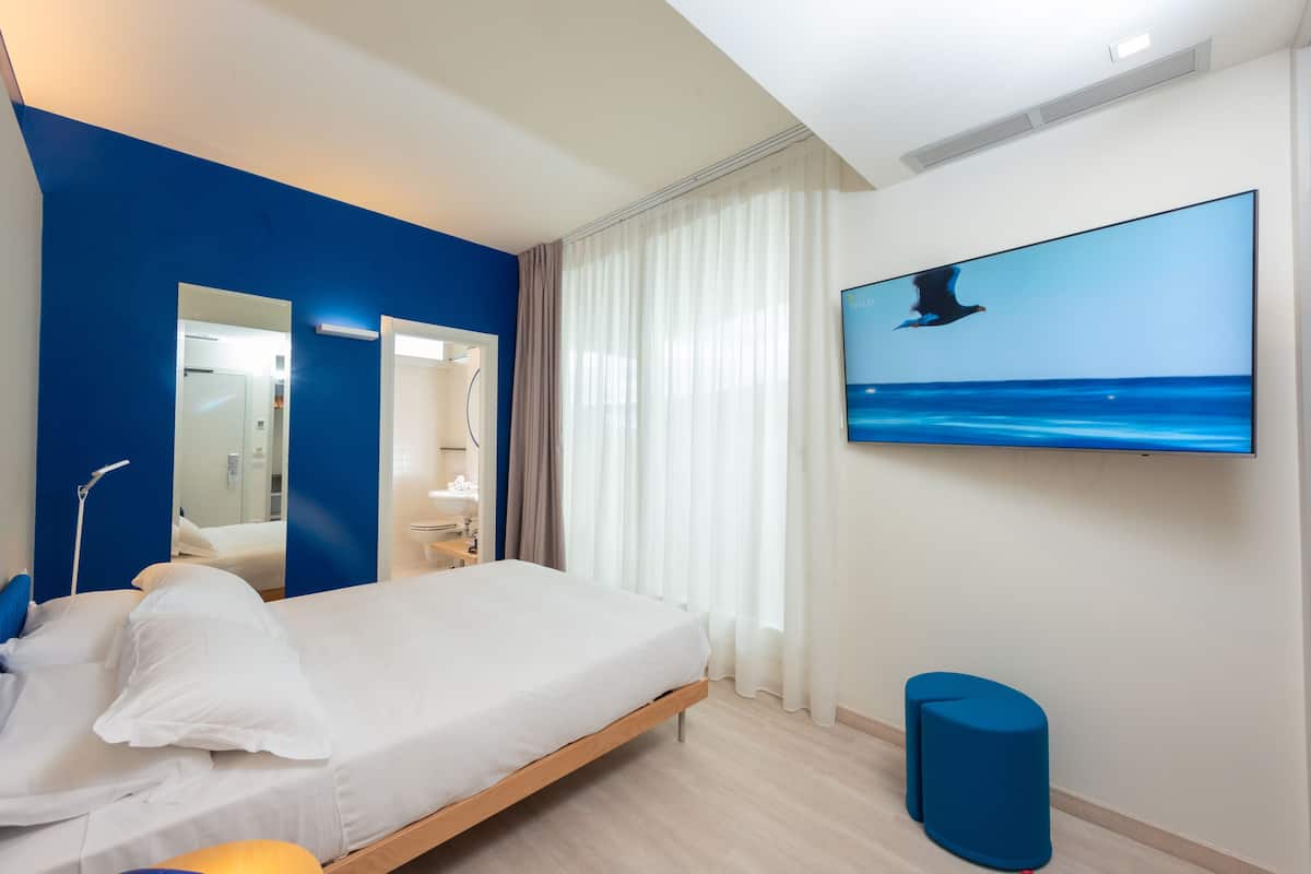 A modern hotel room with a bed, a large window with curtains, a flat-screen TV on the wall, and a blue accent wall.