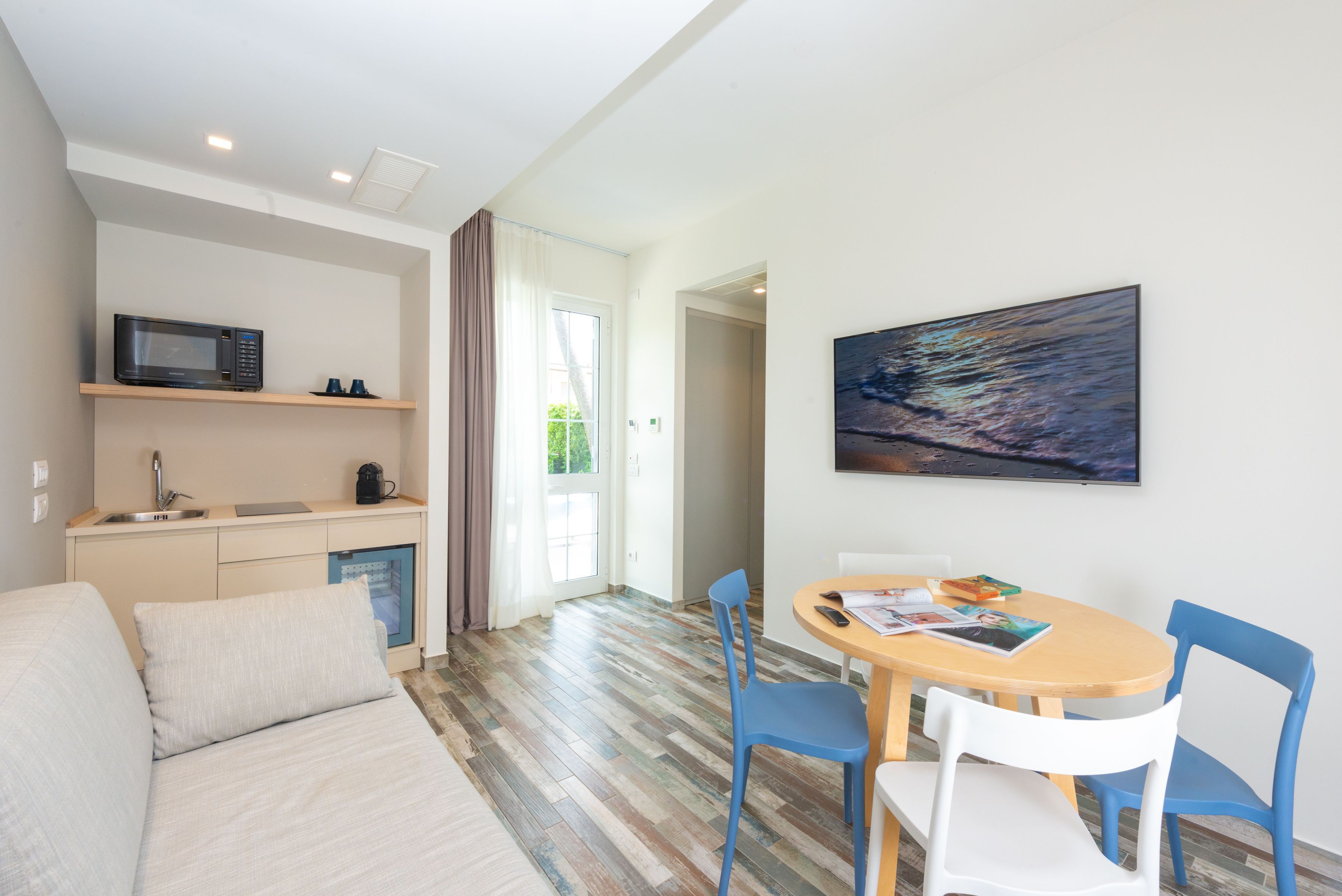 A modern living room with a sofa, a dining table with chairs, a kitchen area with a microwave, and a large painting on the wall.