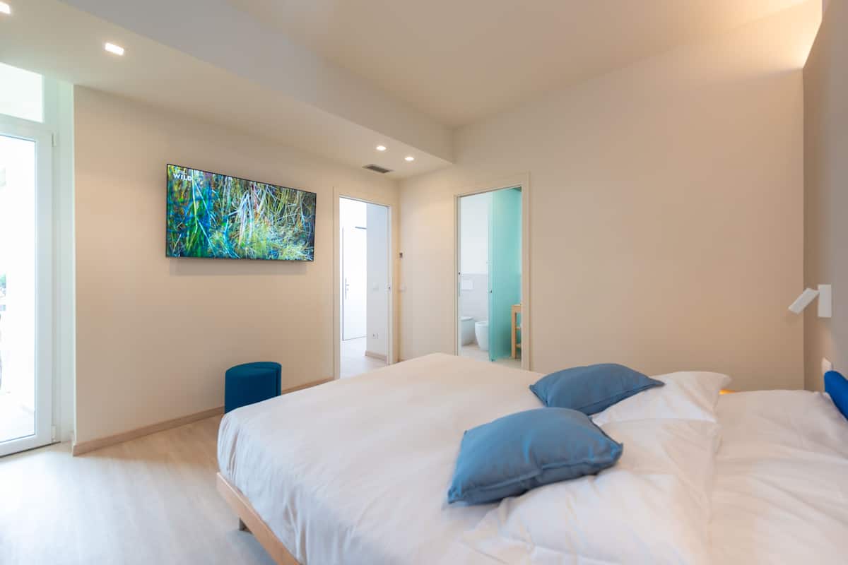 A modern bedroom with a large bed, a painting on the wall, and a view of the bathroom through a mirror.