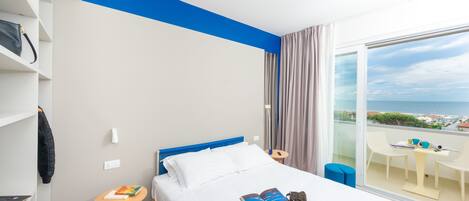 A modern hotel room with a large bed, a small table, a chair, and a balcony with a view.