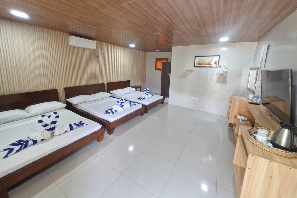 A hotel room with four beds, a wooden ceiling, a flat-screen TV, and a small wooden table with a kettle and cups.