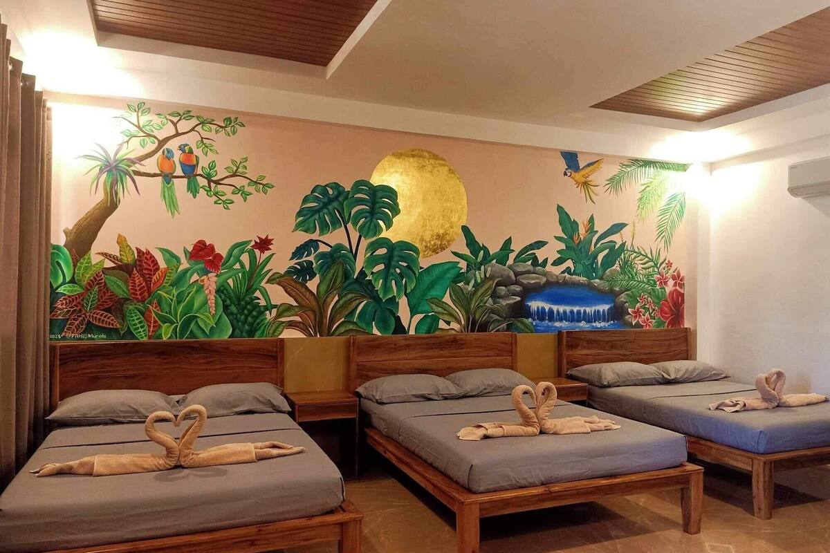A hotel room with three wooden beds, each with a grey mattress and pillows. The room features a large mural on the wall depicting tropical foliage and a waterfall.