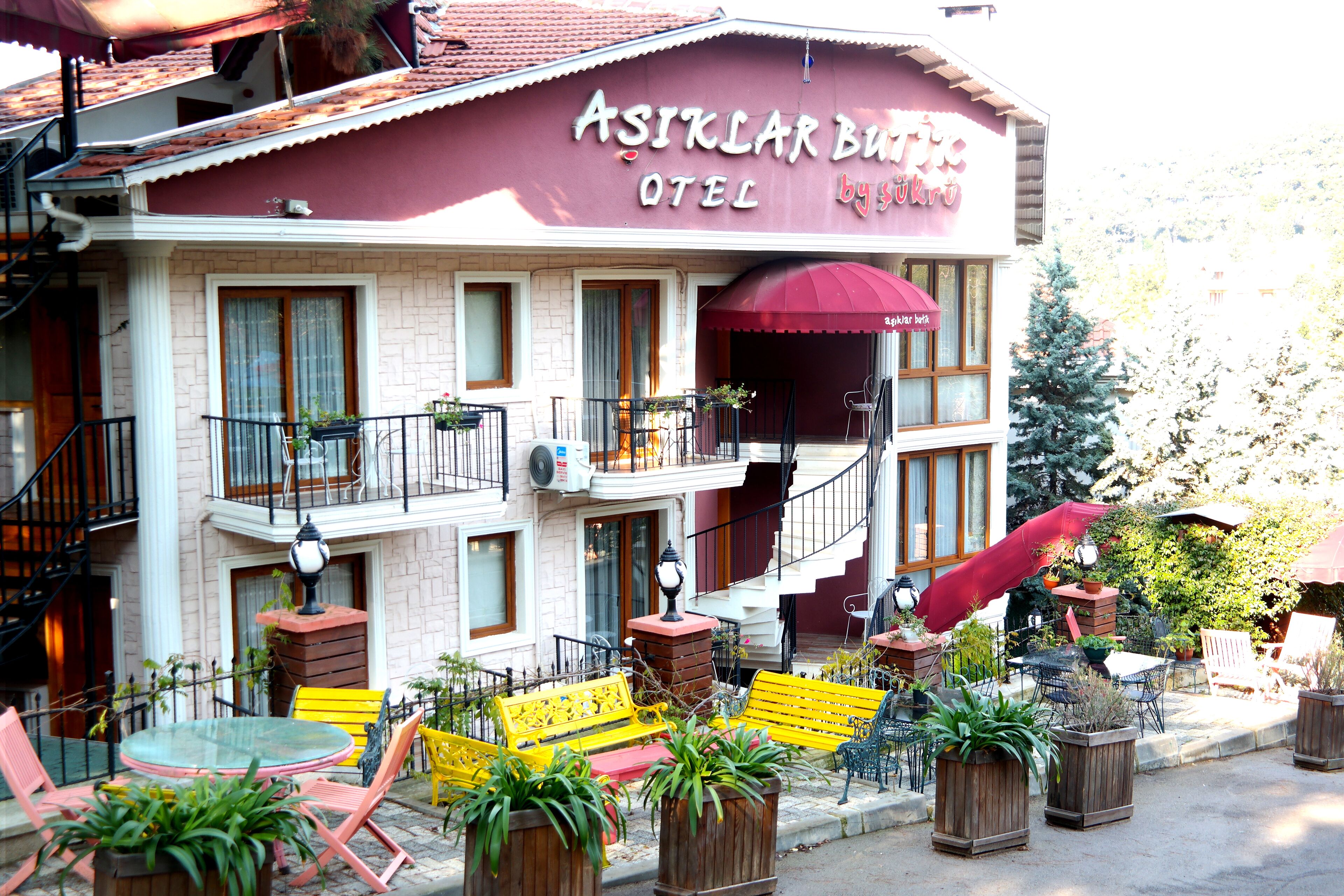 A building with a sign reading "Asiklar Bula Hotel by Sükür". It has a balcony with chairs and tables, and potted plants around the entrance.