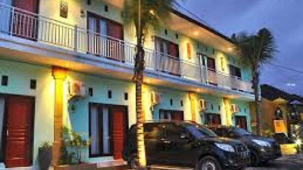 Un hotel de varios pisos con balcones, un coche estacionado al frente y una palmera.